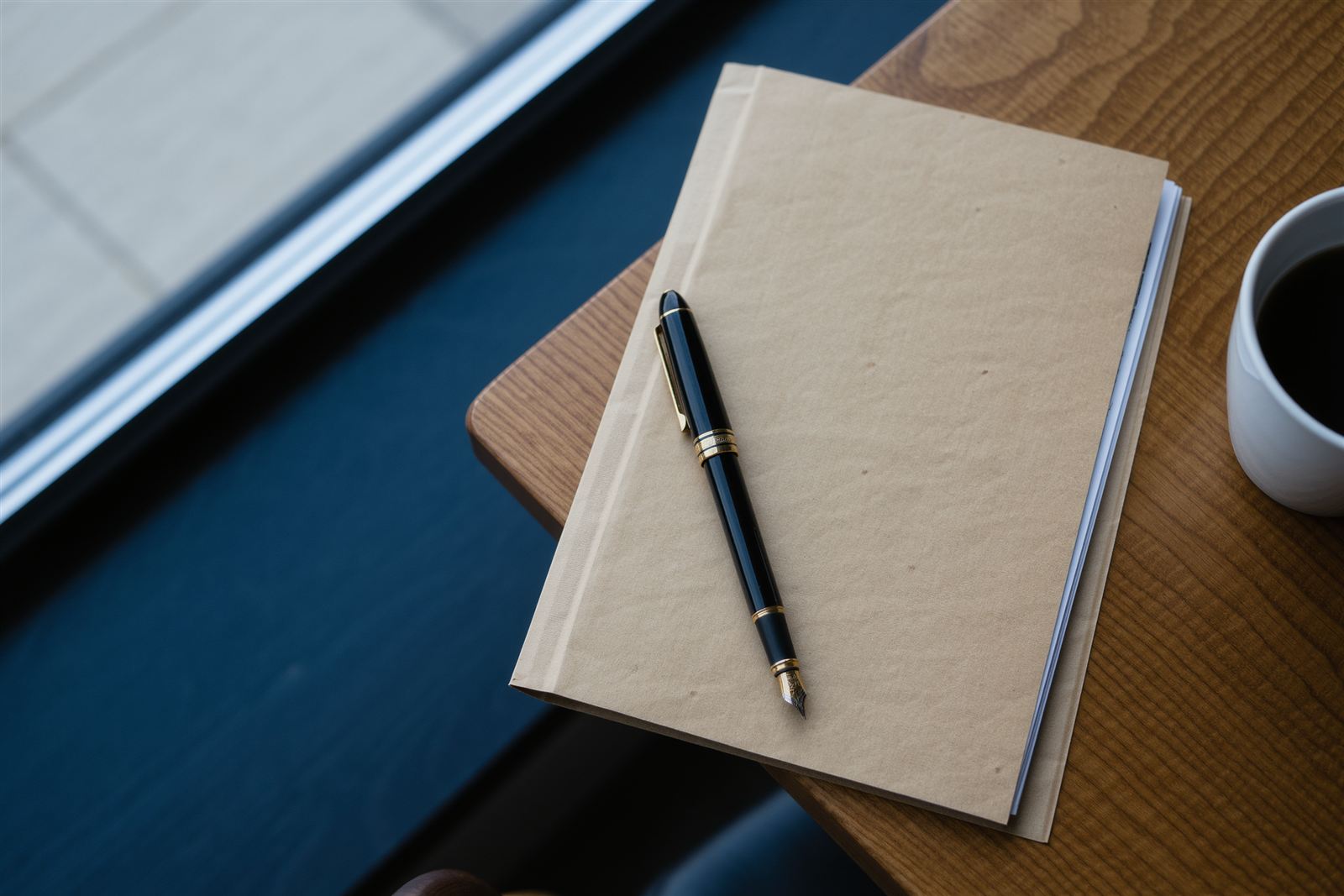 A closed manila folder and a fountain pen on a wooden table beside a window