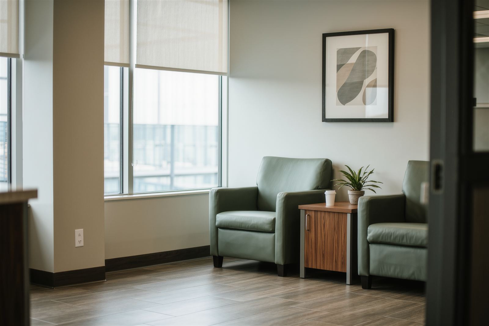 Two green armchairs and a wooden side table with a plant in a bright waiting area